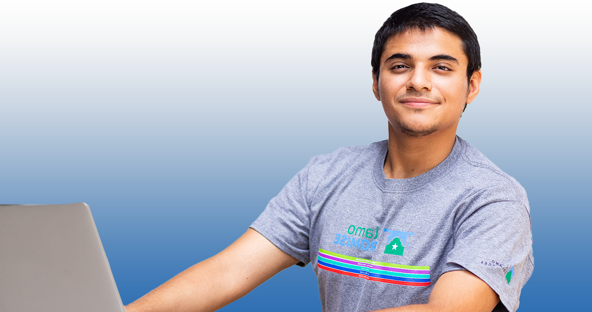 Male student smiling while working on a laptop; student is wearing a grey Alamo Promise shirt with the logo on the front